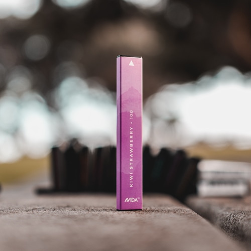 An AVIDA Kiwi Strawberry disposable CBD vape pen standing upright on an outdoor surface, with a blurred background that highlights the product's sleek design.