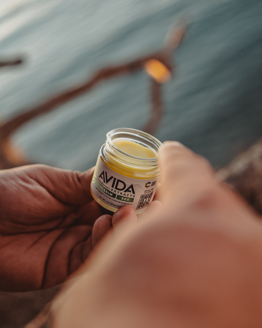 AVIDA 500mg CBD balm jar being opened, showcasing the smooth balm texture, with a natural outdoor setting in the background.