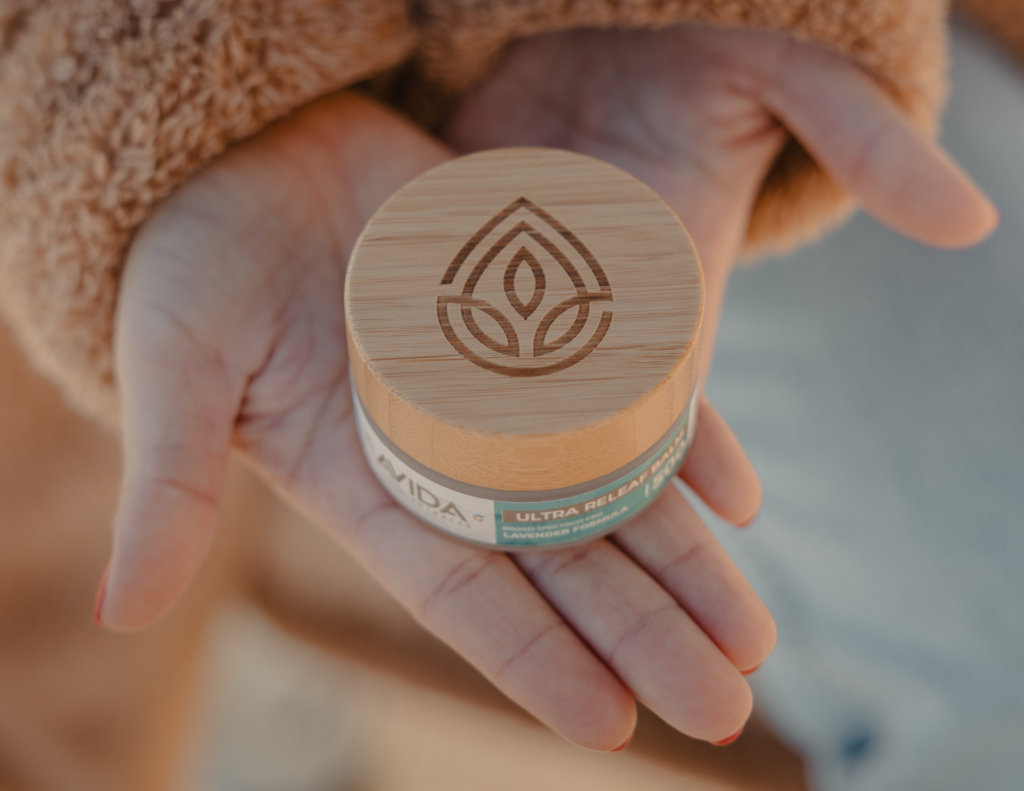 AVIDA Ultra Relief 1000mg CBD balm in a glass jar with a wooden cap with an engraved Avida logo, displayed in an open hand, featuring a broad-spectrum formula with lavender, designed for topical relief.