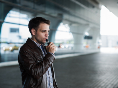 Man with hair and black jacket standing outside airport terminal vaping.
